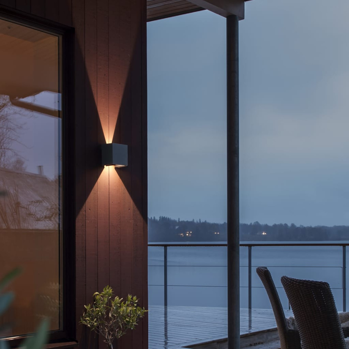 Scatto esterno di una casa. Un'applique è accesa, illuminando la parete in legno. Lo sfondo mostra un lago e alberi.