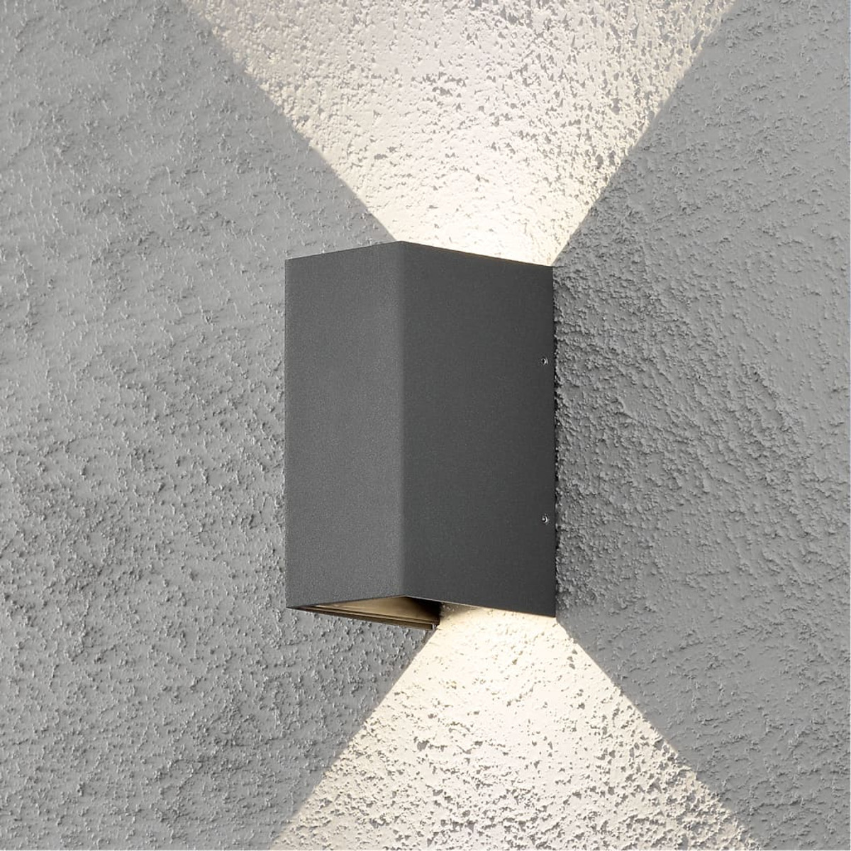 Una lampada da parete rettangolare grigia su un muro grigio strutturato. La luce brilla su e giù, creando un motivo a croce.