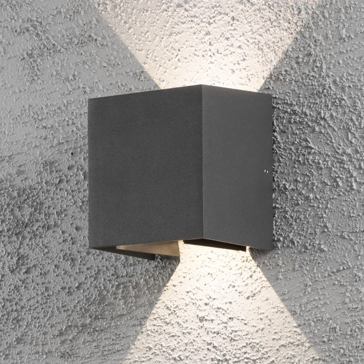 Una lampada da parete esterna quadrata, grigio scuro, con luce che brilla verso l'alto e verso il basso, montata su un muro strutturato grigio chiaro.