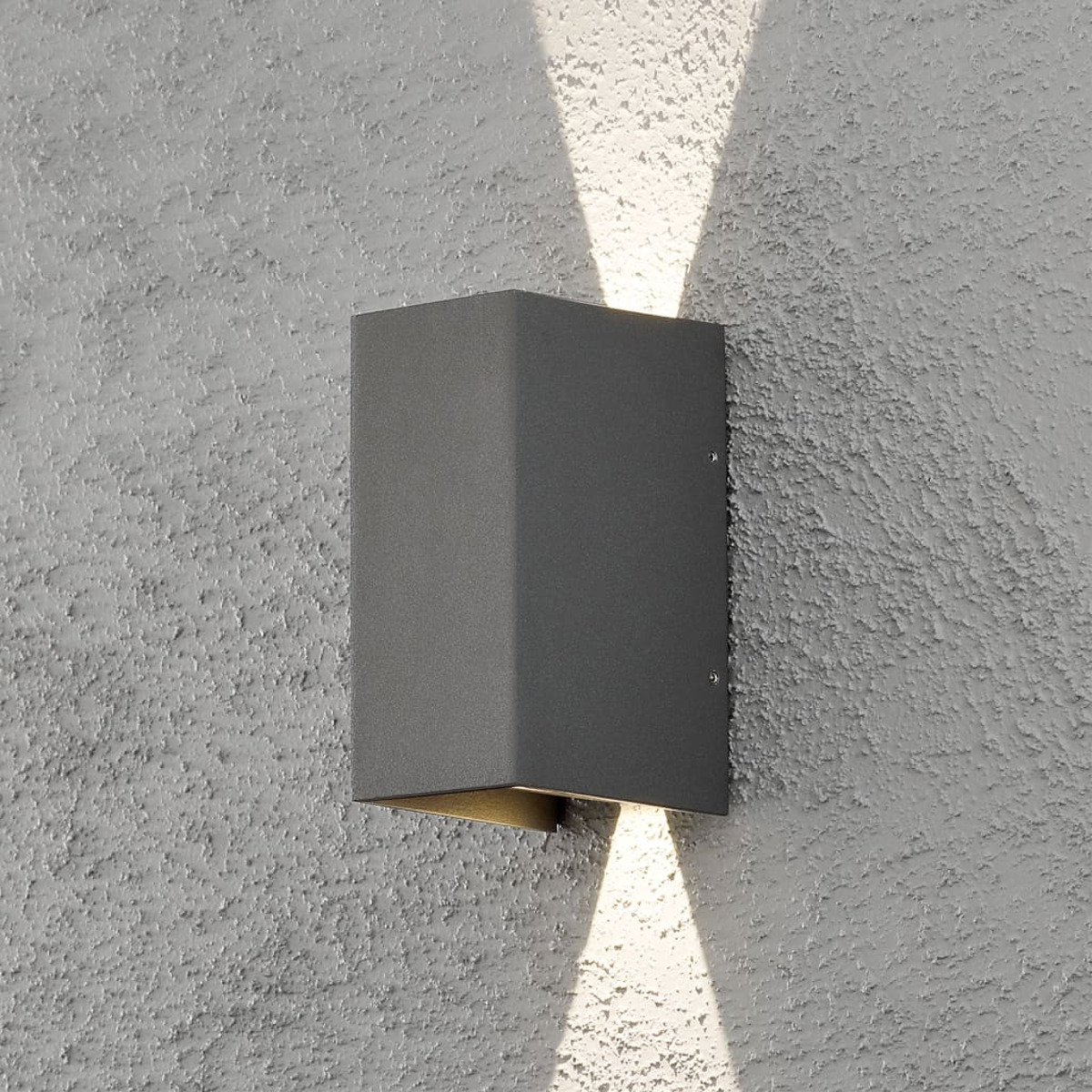 Una lampada da parete esterna rettangolare grigio scuro è montata su un muro grigio chiaro strutturato, che proietta un fascio di luce verso l'alto e verso il basso.