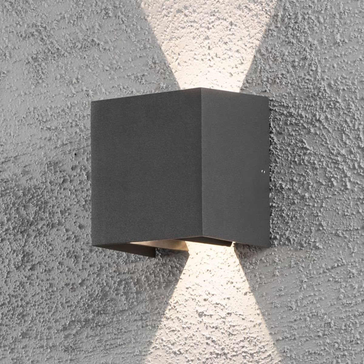 Un apparecchio di illuminazione esterno quadrato grigio scuro è montato su un muro grigio strutturato, che proietta due fasci di luce.