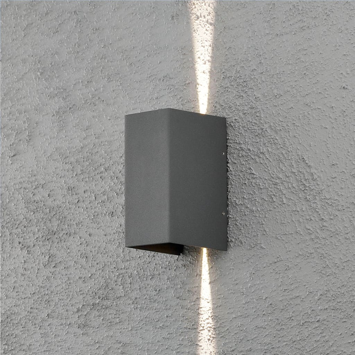 Una lampada grigia a parete emette luce verso l'alto e verso il basso. Il muro è strutturato e verniciato di grigio chiaro.