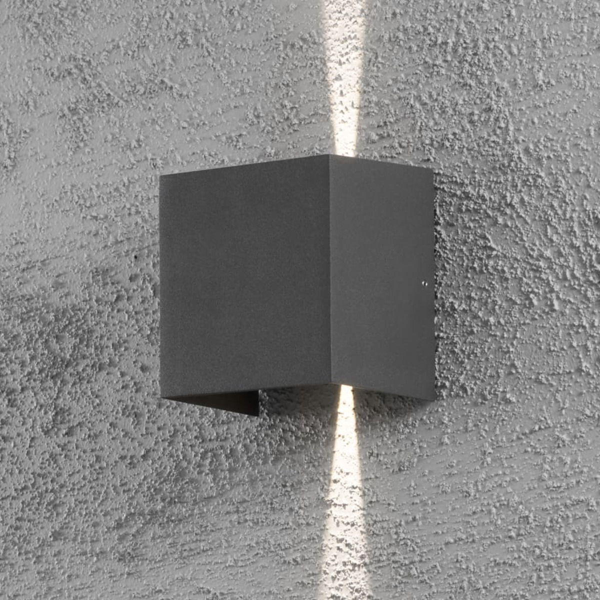 Una lampada da parete quadrata grigio scuro su un muro grigio strutturato. I fasci di luce vanno su e giù. Il muro ha una consistenza ruvida, simile allo stucco.