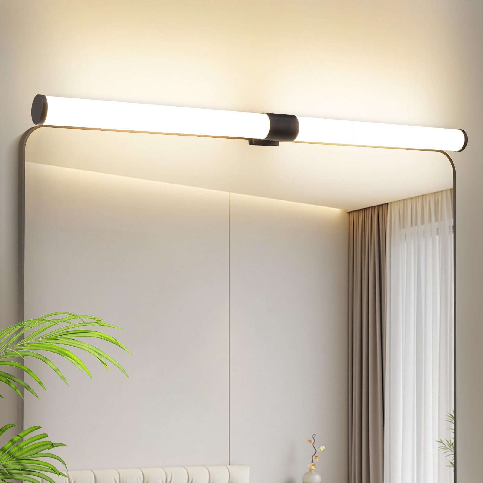 Uno specchio illuminato con un apparecchio nero sopra. Felci e tende trasparenti ai lati. Lo sfondo è beige.