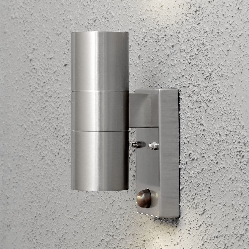 Una moderna lampada da parete esterna in metallo è montata su un muro grigio strutturato. Ha un design cilindrico con sensore.