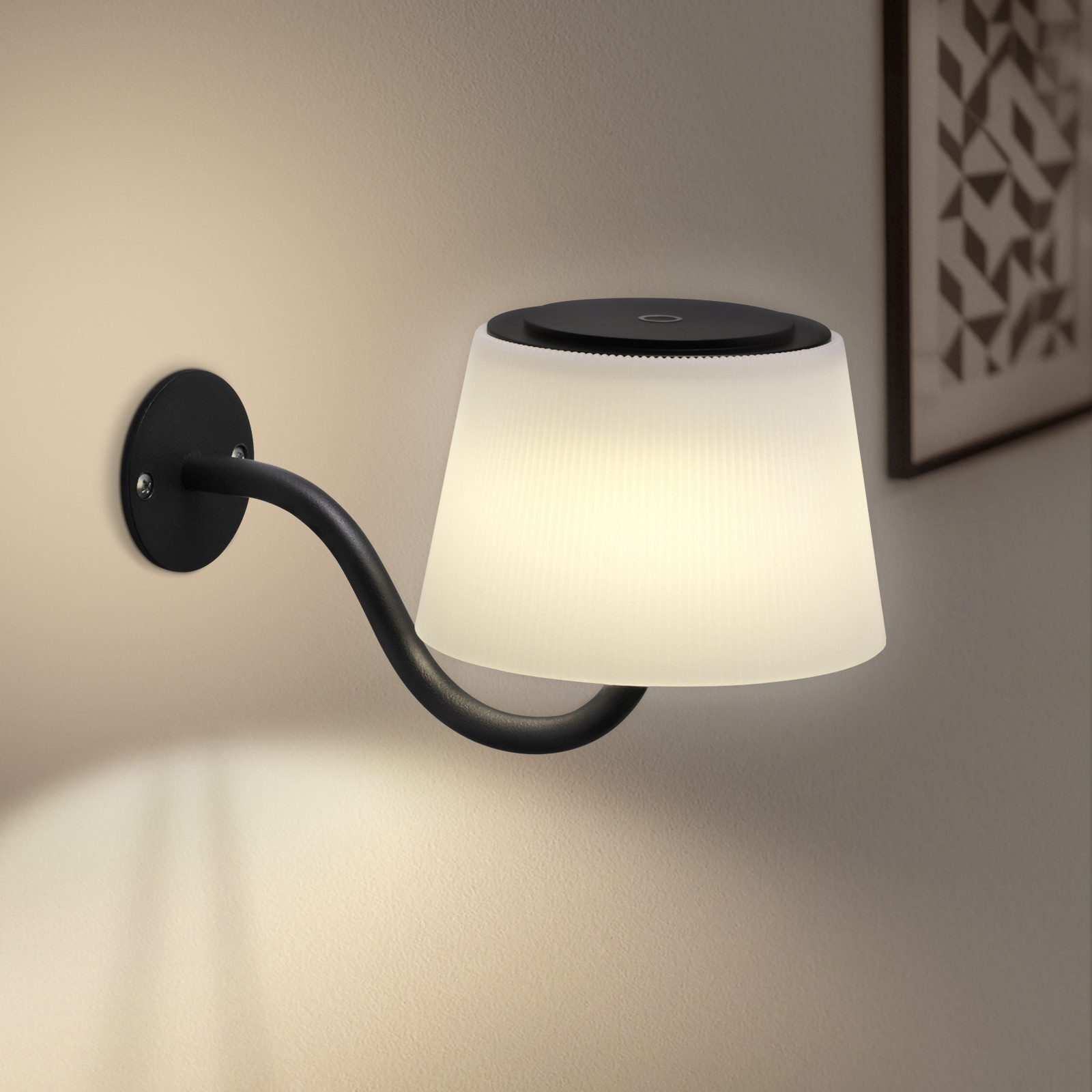 Lampada da parete nera con paralume bianco. Montata su un muro beige. Un'opera d'arte incorniciata sullo sfondo.