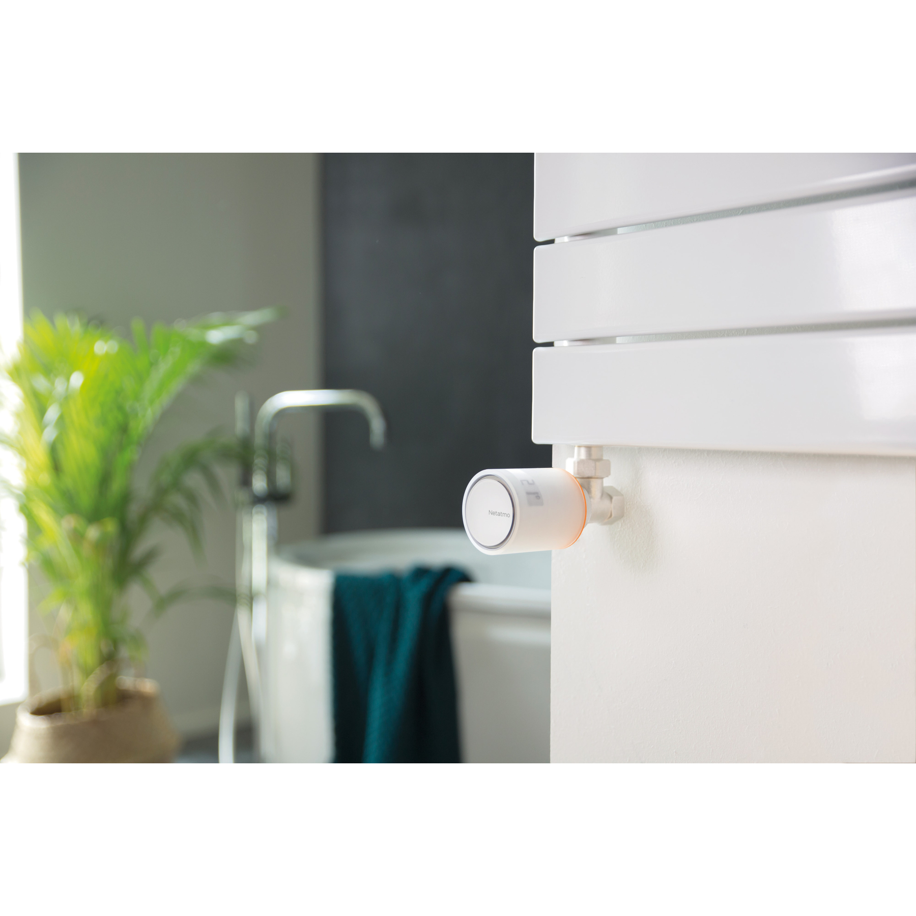 A bathroom scene with a smart thermostat on a white radiator. A bathtub, green plant, and towel are visible.