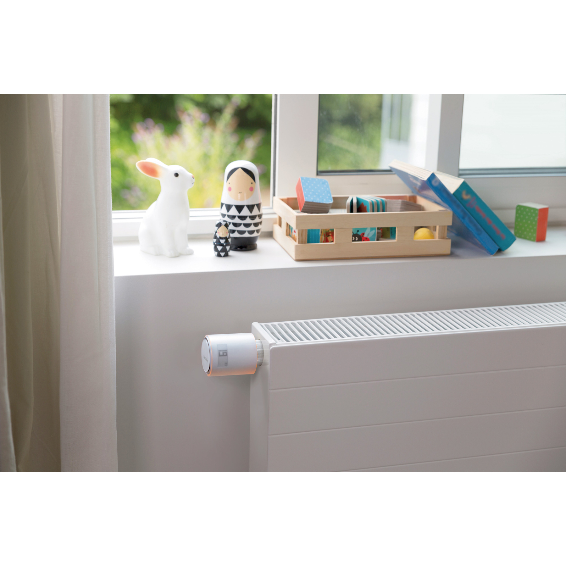 A window sill with toys and books. A radiator has a thermostat. A curtain is on the left.
