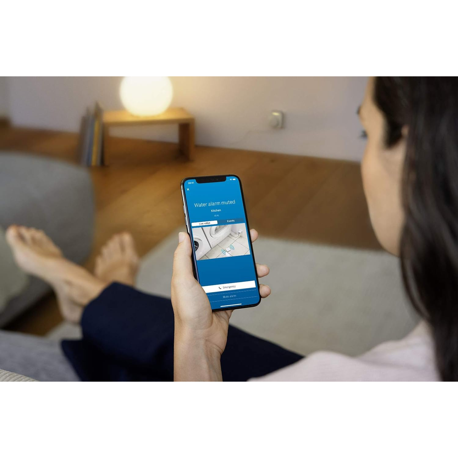 Osoba trzyma telefon z wyciszonym alarmem wody. Scena obejmuje lampę, kanapę i nogi osoby.