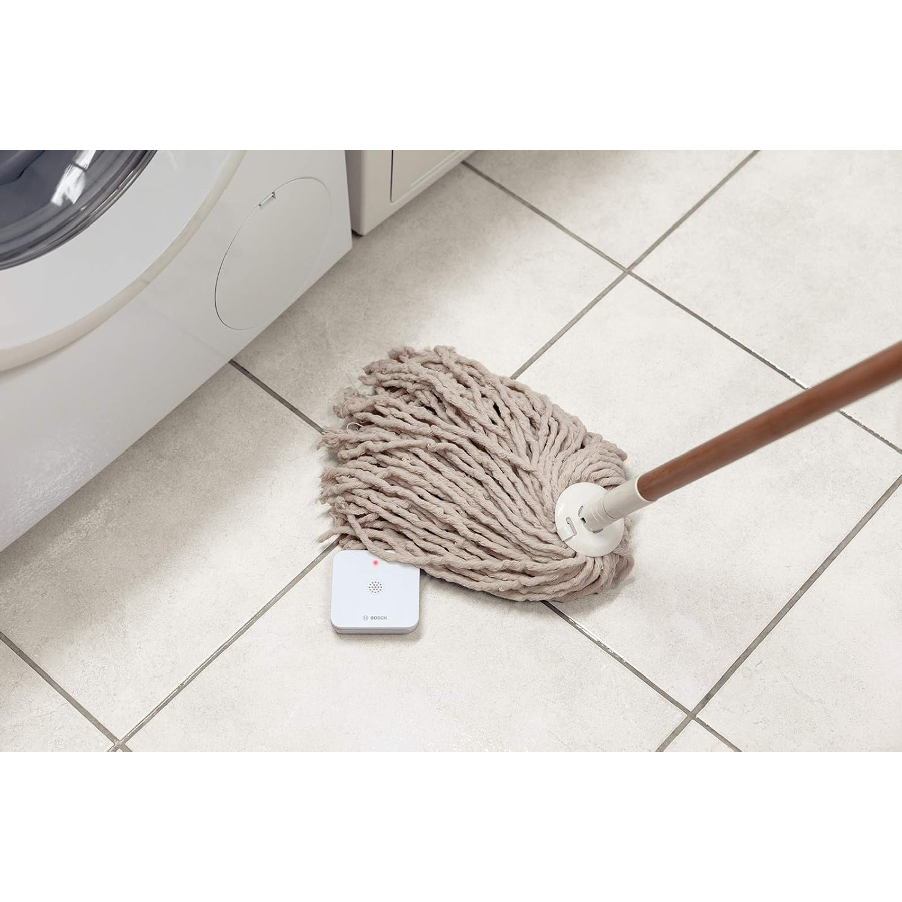 Mop z brązowym uchwytem leży na kafelkach obok pralki. Biały czujnik leży na podłodze.