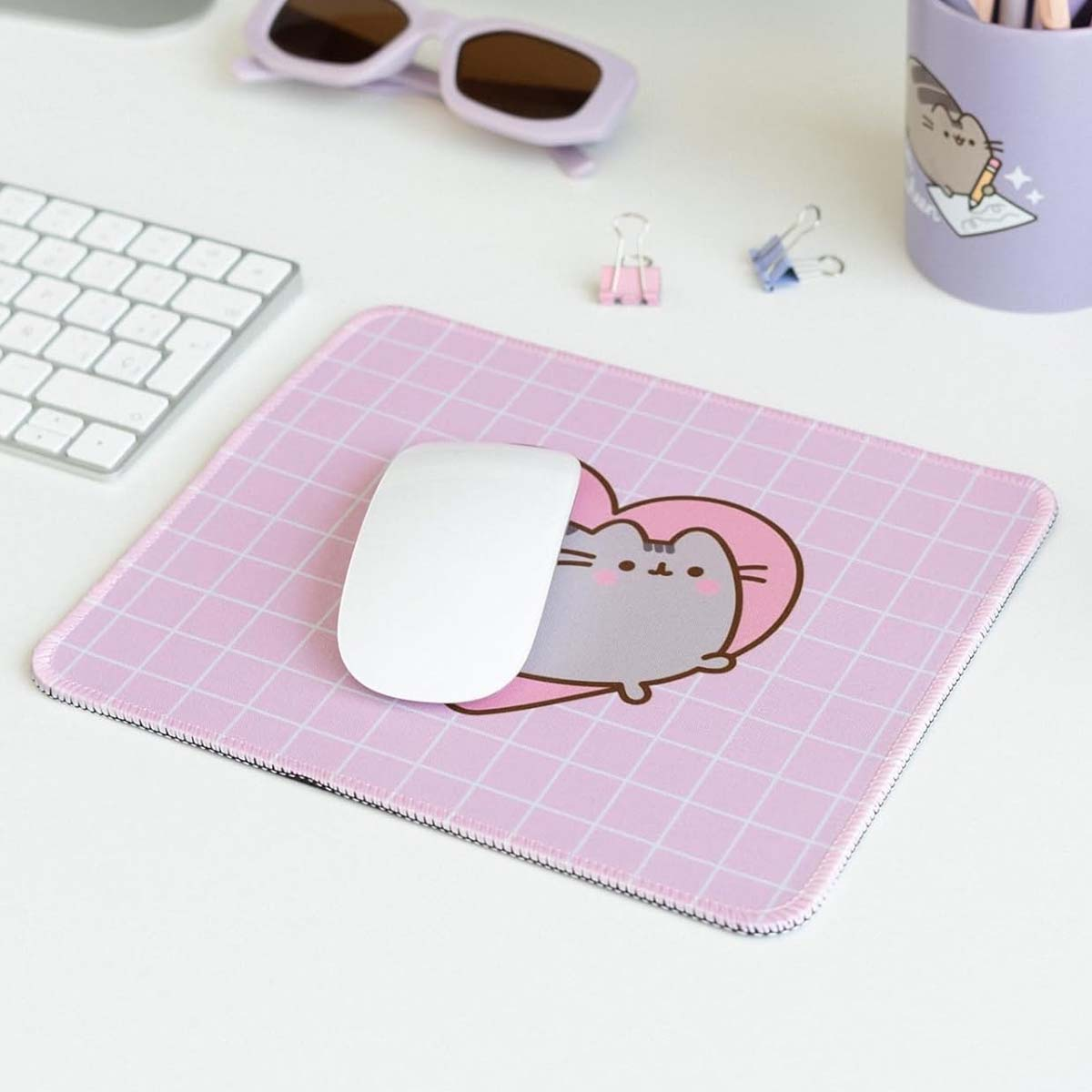 Un mousepad rosa con un design a gatto, un mouse bianco, una tastiera, occhiali da sole viola, clip e un portapenne sono su una scrivania bianca.