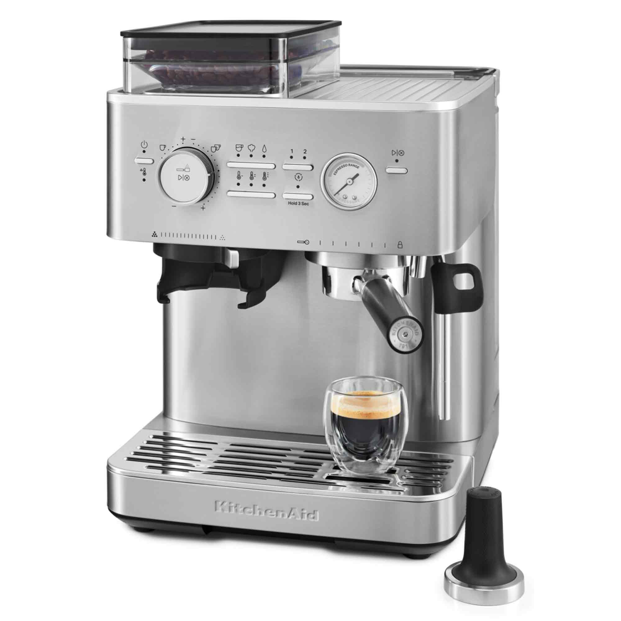 Eine silberne KitchenAid Espressomaschine mit einem Glas Kaffee und einem schwarzen Tamper. Kaffeebohnen sind oben sichtbar.