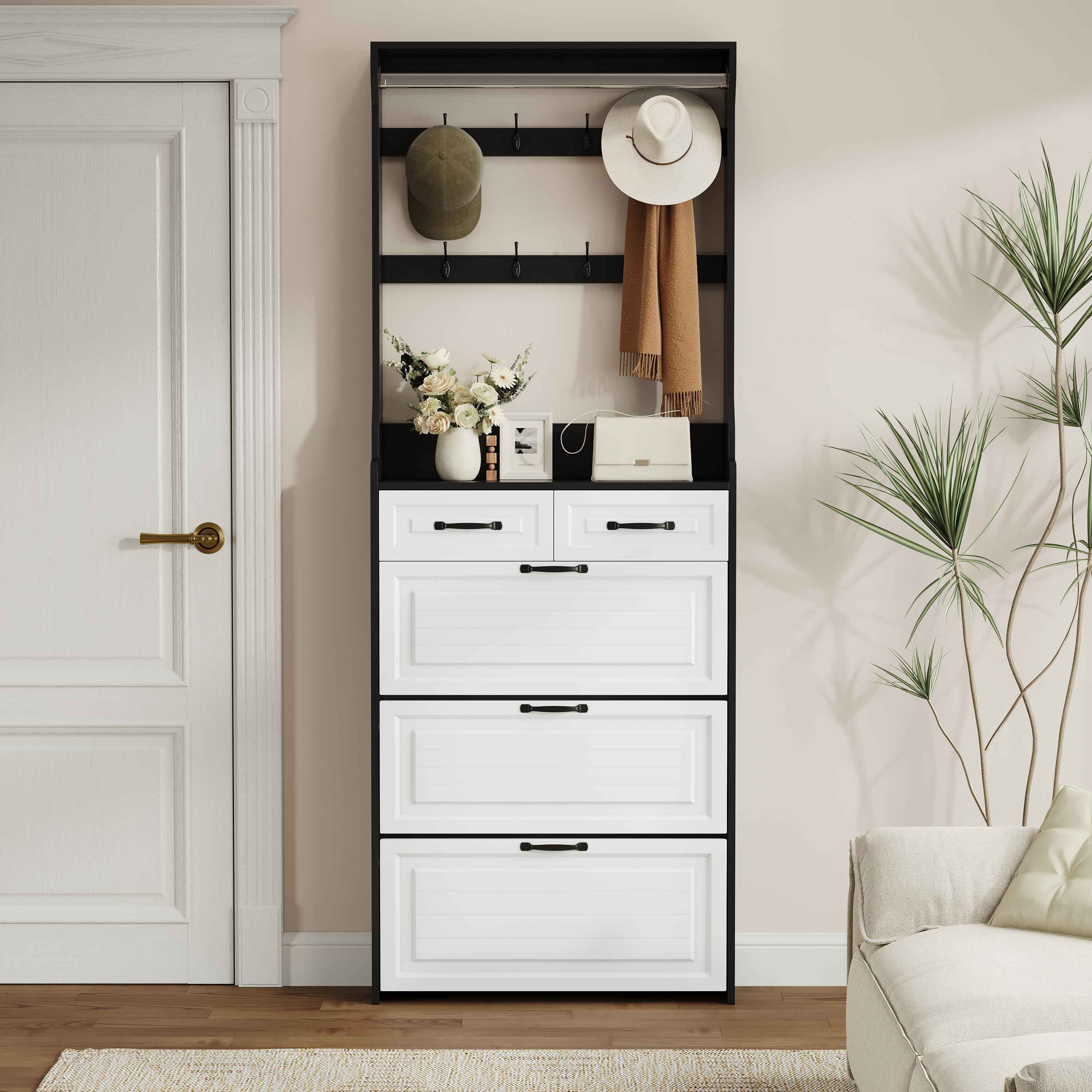 Eine weiße Tür, ein schwarz-weißer Schrank, ein Hut, ein Schal und Blumen. Im Hintergrund befinden sich ein Sofa und eine Pflanze.