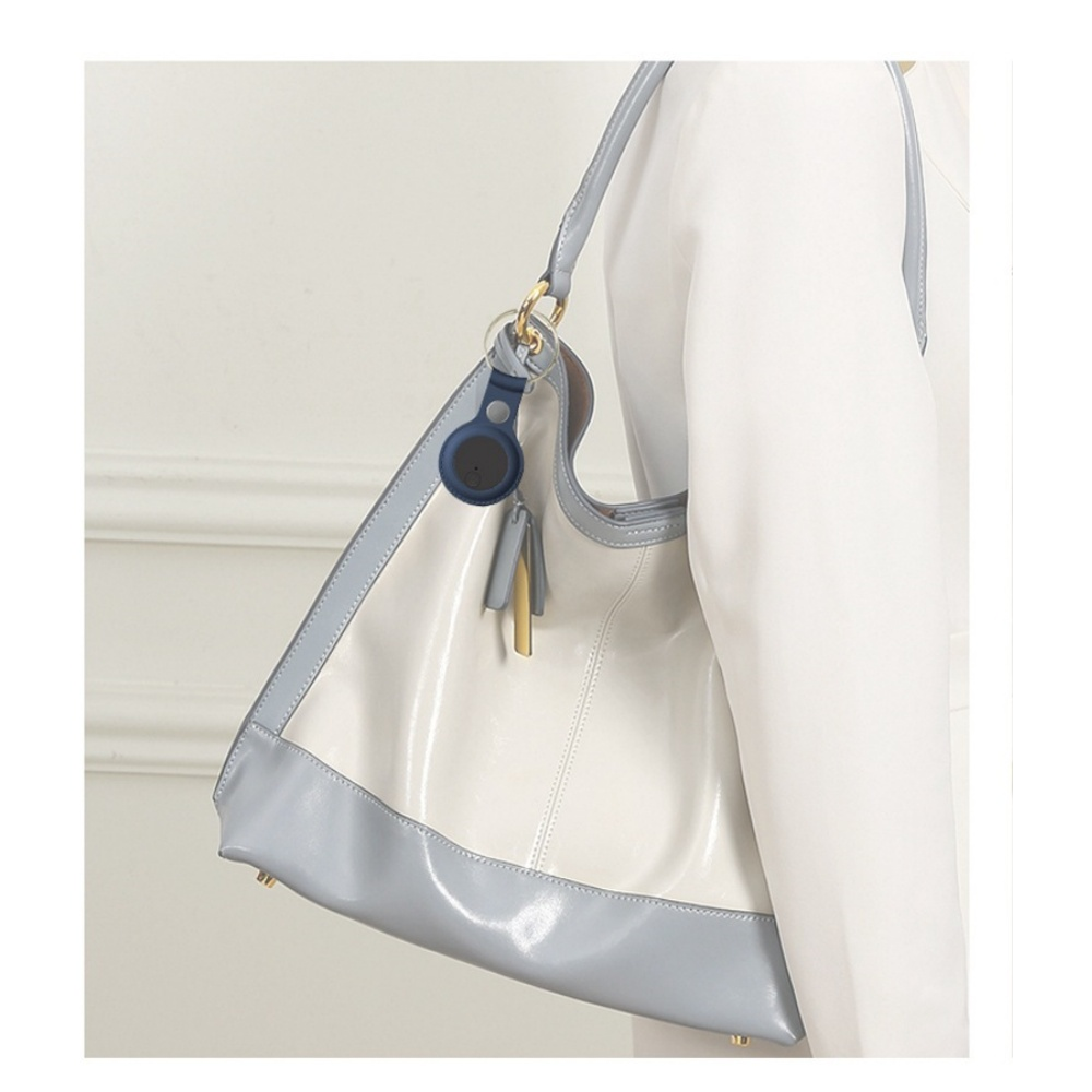 Eine glänzende, weiß-hellblaue Handtasche wird von einer Person gehalten, die einen weißen Blazer trägt. Ein blauer Tracker ist am Griff befestigt.