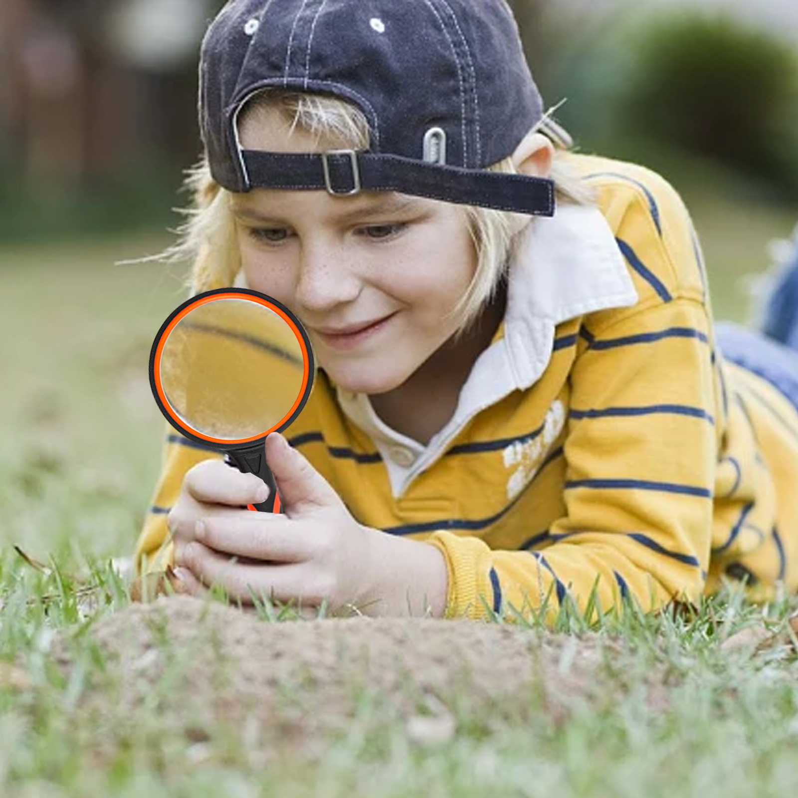 Ein Junge mit einer Lupe untersucht etwas auf dem Boden. Er trägt eine Kappe und ein gelb gestreiftes Hemd und liegt im Gras.