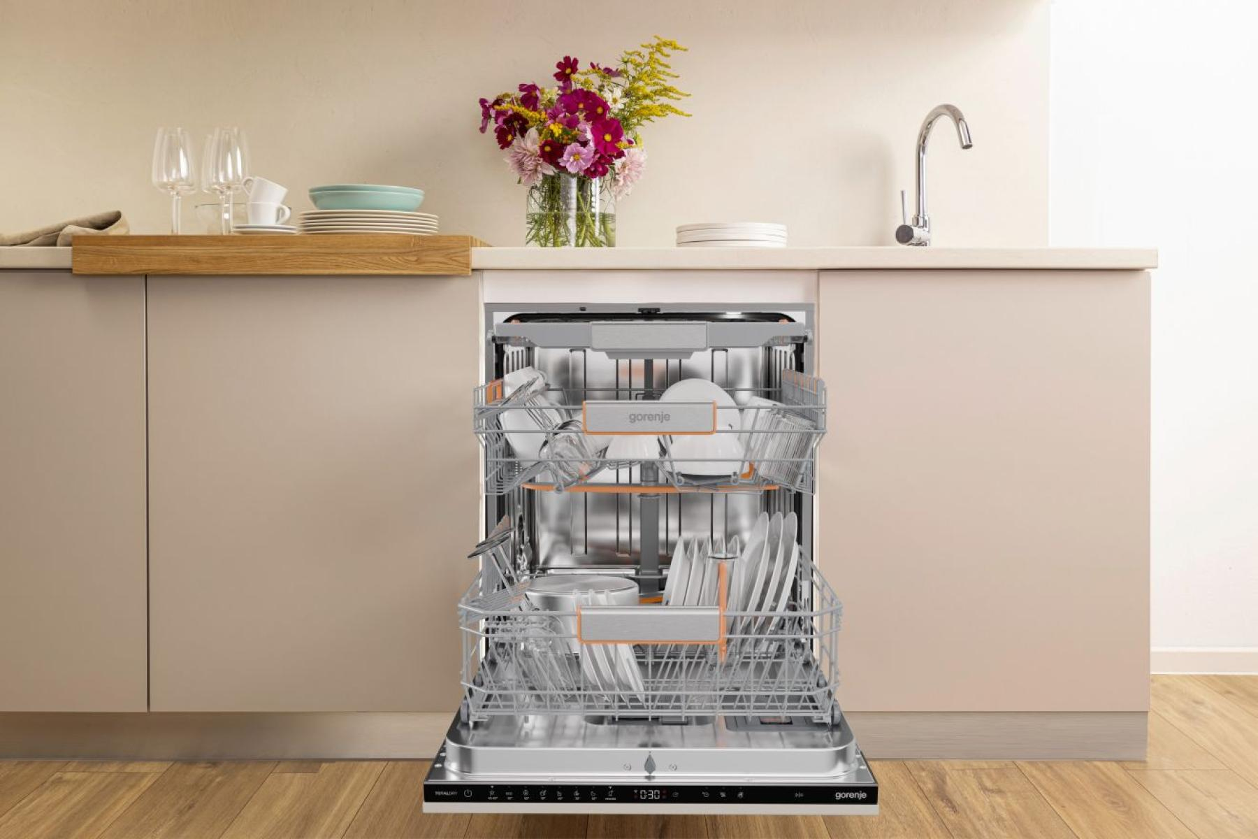 Lavastoviglie aperta con piatti. Cucina con piano di lavoro, fiori e rubinetto. Mobili beige e pavimento in legno. Marchio Gorenje.