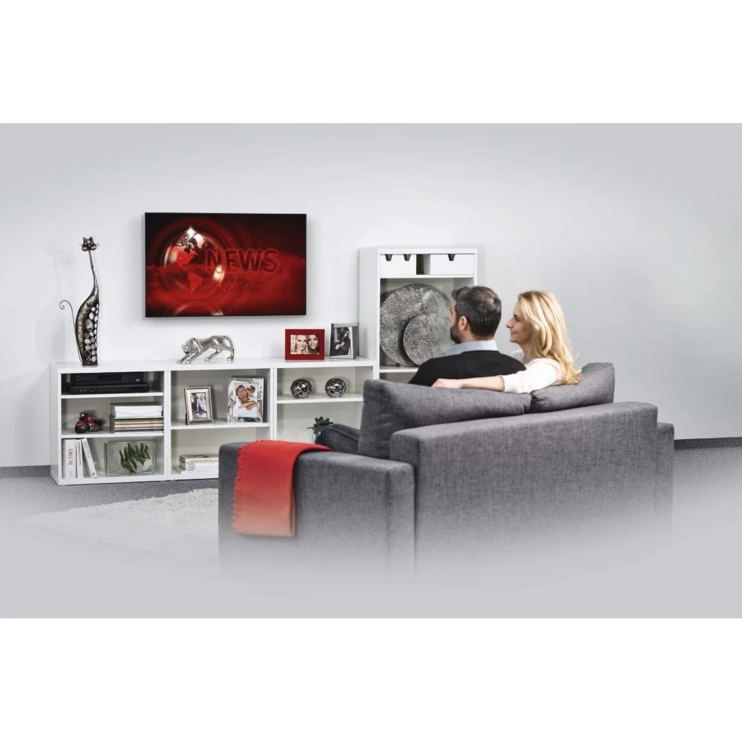Ein Paar sitzt auf einem grauen Sofa und schaut fern. Ein roter Überwurf liegt auf dem Sofa. Ein weißes Regal hält Deko und ein Fernseher zeigt die Nachrichten.