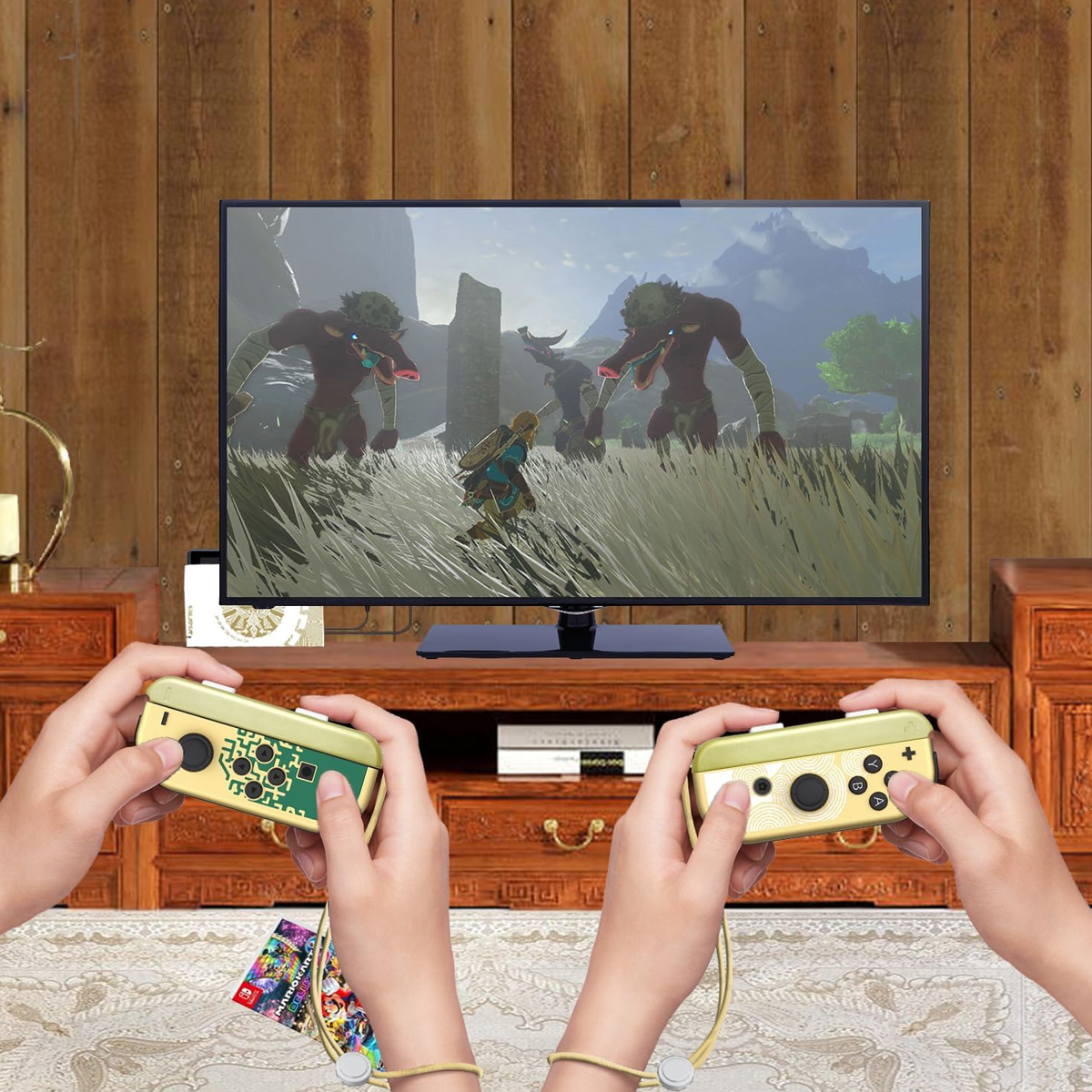 Zwei Hände halten Controller und spielen ein Spiel, das auf einem Fernseher angezeigt wird. Das Spiel zeigt Charaktere in einem Feld mit Bergen im Hintergrund.