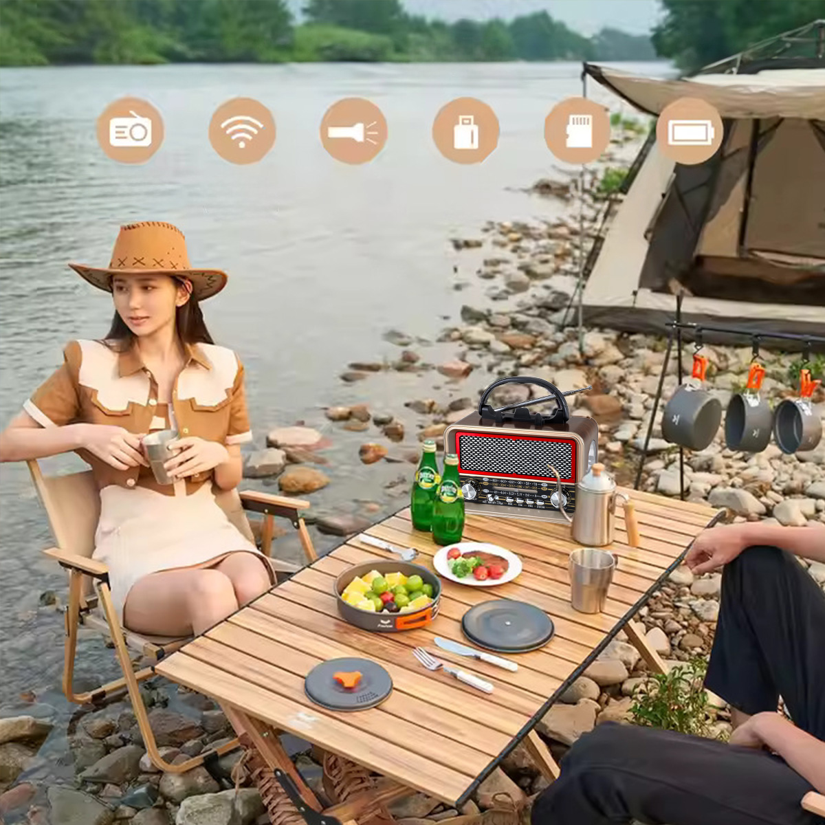 Kobieta w kapeluszu siedzi przy stole z jedzeniem. Radio, napoje i sprzęt kempingowy są w pobliżu. Namiot i rzeka w tle.