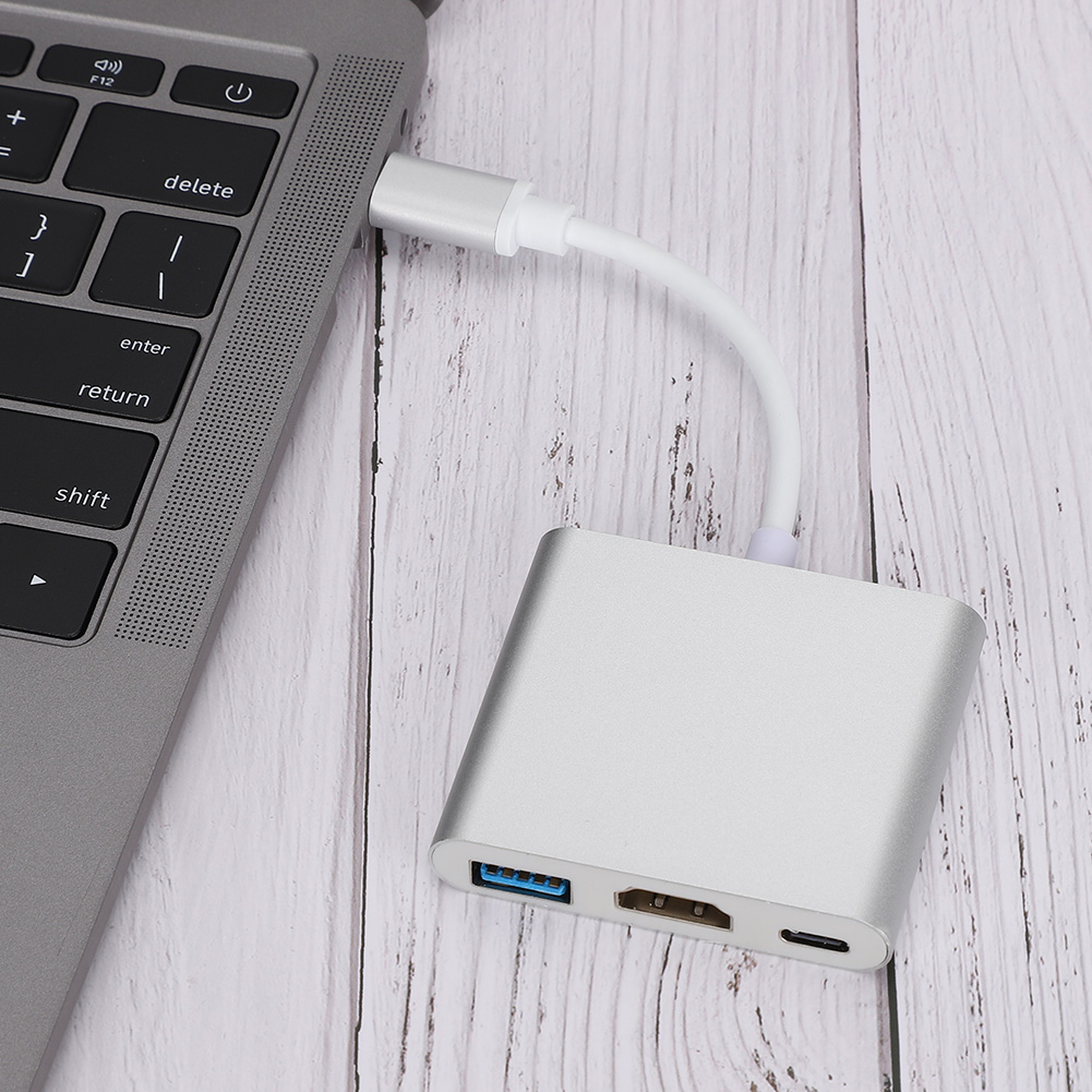Ein silberner USB-C-Hub, der mit einem Laptop verbunden ist. Der Hub hat USB-, HDMI- und USB-C-Anschlüsse. Der Hintergrund ist eine Holzoberfläche.