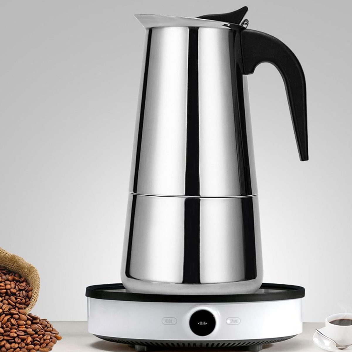 Ein Edelstahl-Kaffeebereiter steht auf einem weißen Sockel. Kaffeebohnen sind in einem Beutel und eine Tasse Kaffee auf einem Tisch.