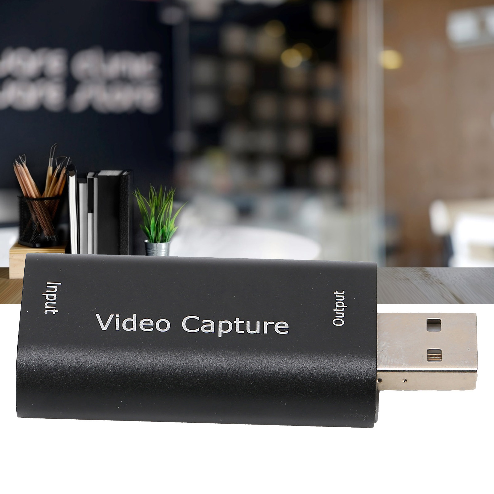 Ein schwarzes Videoaufnahmegerät mit USB-Anschluss. Text auf dem Gerät: Input, Video Capture, Output. Hintergrund: Büroumgebung.