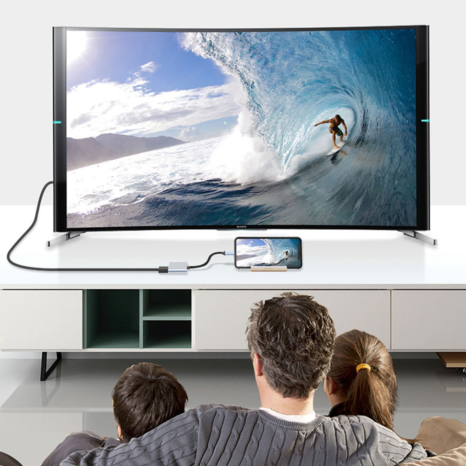 Ein gebogener Fernseher zeigt einen Surfer auf einer Welle. Ein Telefon zeigt dasselbe Bild, das mit dem Fernseher verbunden ist. Eine Familie schaut zu.