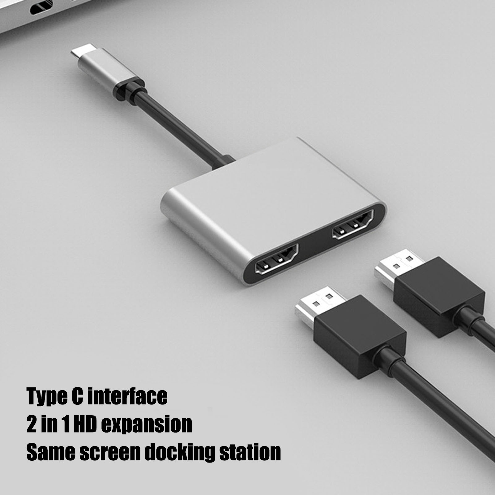 Ein silberner USB-C-Hub mit zwei HDMI-Anschlüssen und zwei schwarzen HDMI-Kabeln auf grauer Oberfläche.
