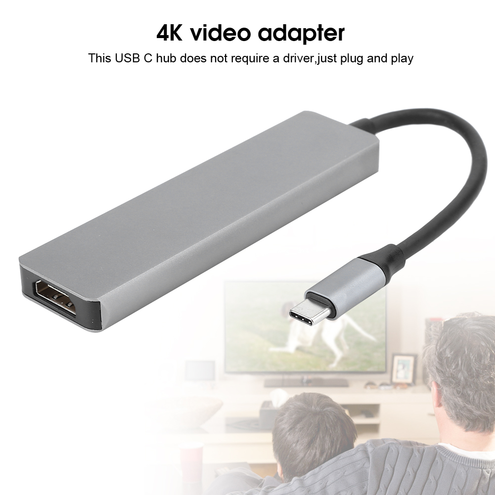 Ein silberner USB-C-Hub mit schwarzem Kabel. Der Hub ist mit einem USB-C-Stecker verbunden. Im verschwommenen Hintergrund sehen zwei Personen fern.