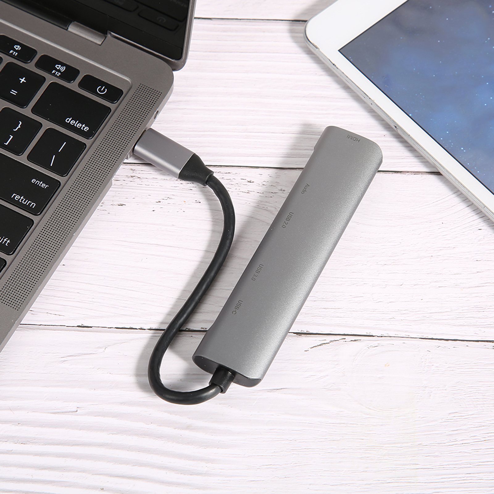 Laptop mit grauem USB-Hub verbunden. Ein Tablet ist im Hintergrund. Alles auf einer weißen Holzoberfläche.