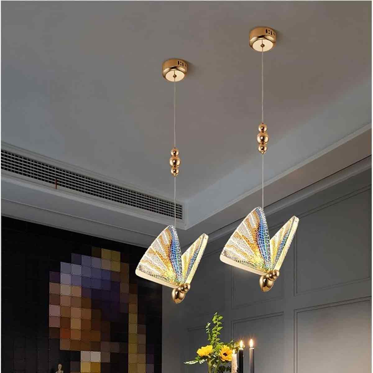 Zwei schmetterlingsförmige Pendelleuchten. Jede hat Mosaikflügel, goldene Akzente und hängt von der Decke. Blumen und Kerzen im Vordergrund.
