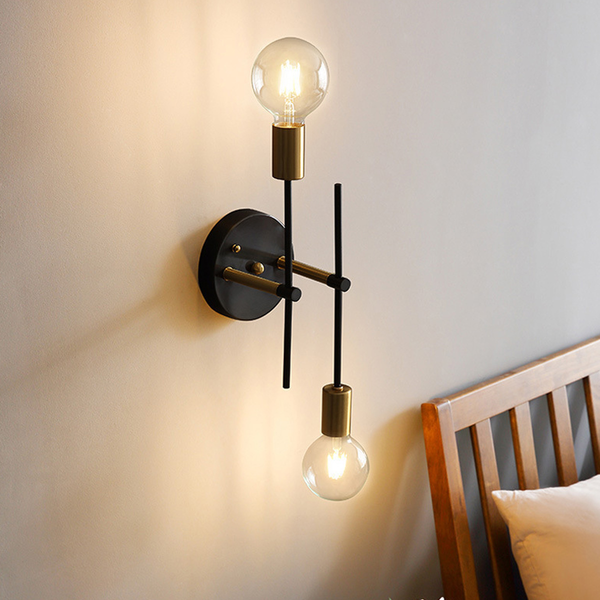 Eine moderne Wandlampe mit zwei brennenden Glühbirnen. Sie ist schwarz und goldfarben, an einer beigen Wand montiert, mit einem Bettrahmen und Kissen.