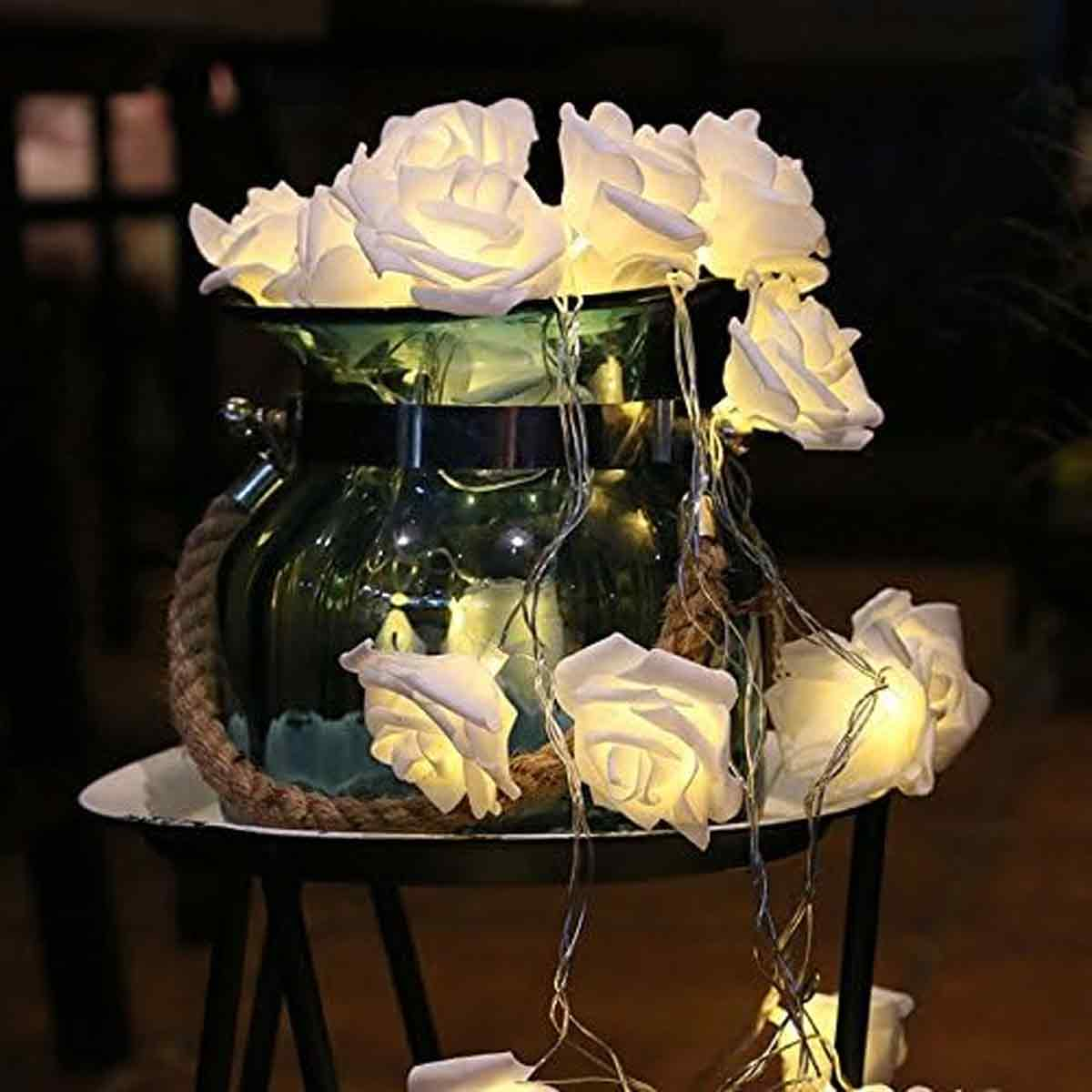 Eine Glasvase mit beleuchteten weißen Rosenlichtern. Die Vase steht auf einem kleinen Tisch mit einem Seil um sie herum. Der Hintergrund ist dunkel.