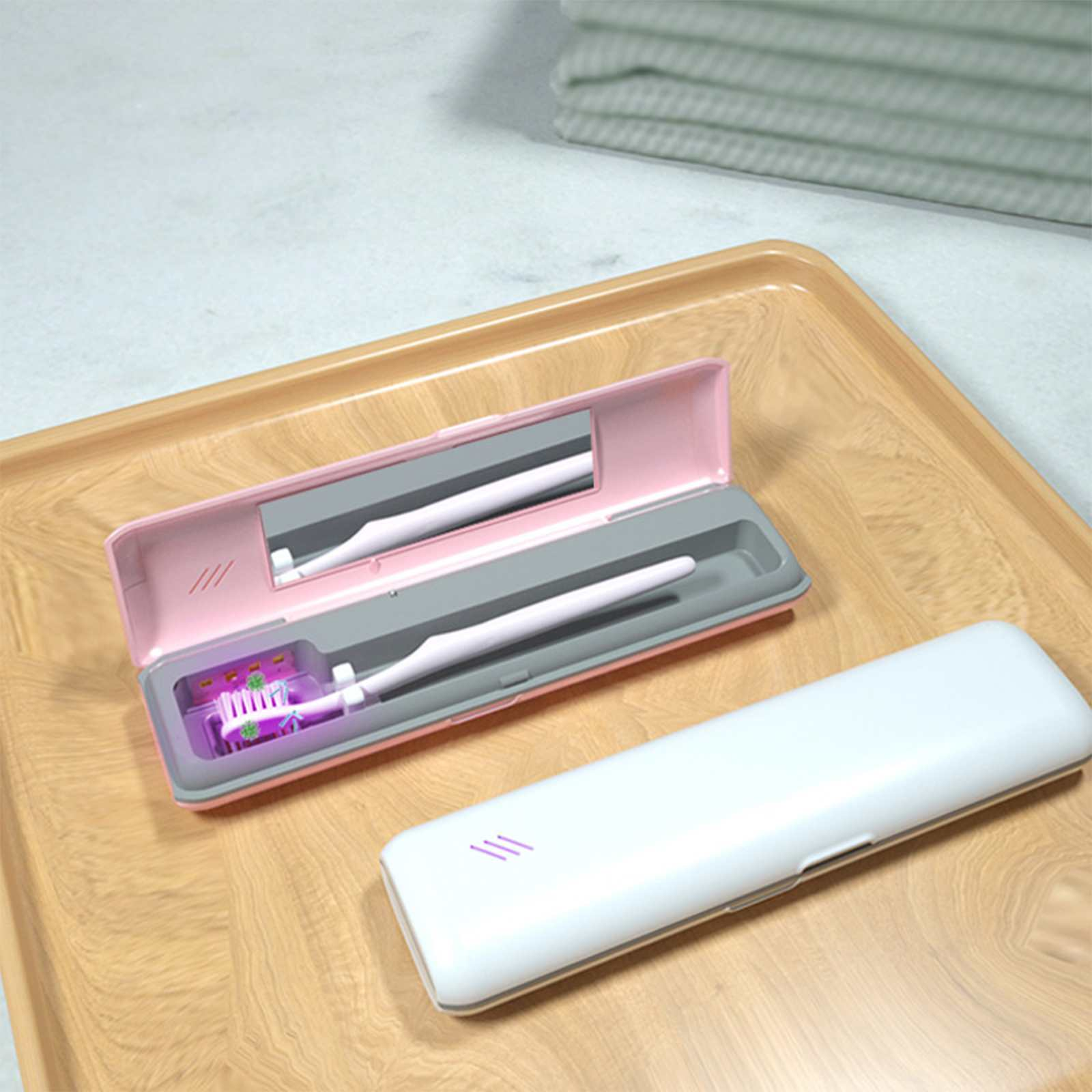 Eine rosa Zahnbürstenhülle ist geöffnet, mit einer Zahnbürste darin. Eine geschlossene weiße Hülle liegt daneben auf einem Holztablett. Ein Handtuch ist im Hintergrund.