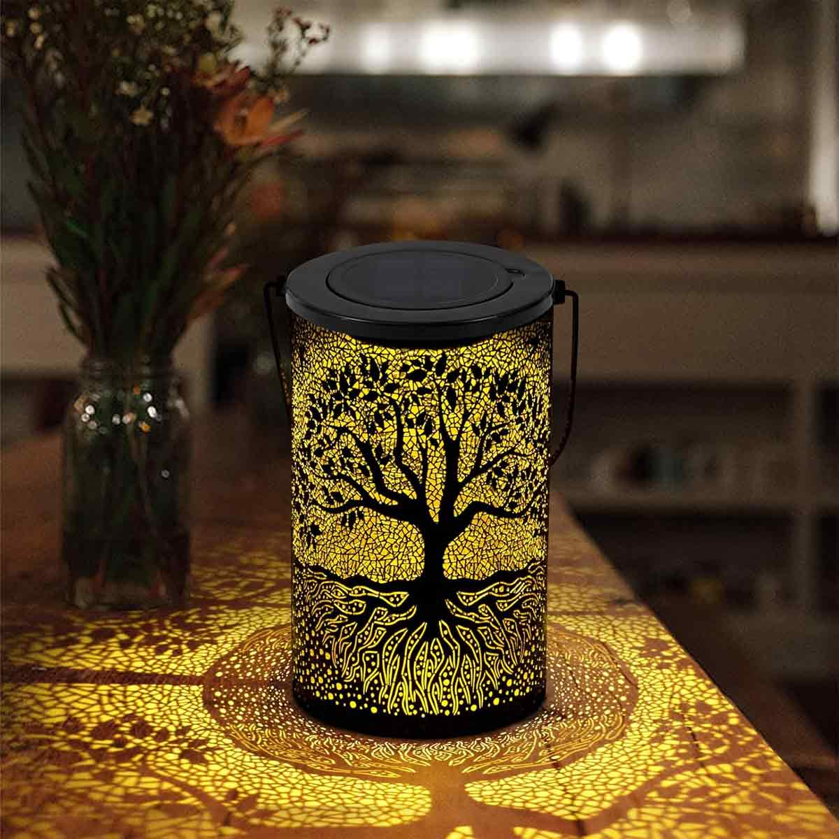 Eine baumförmige Laterne strahlt gelbes Licht aus und wirft einen Baumschatten auf den Tisch. Eine Vase mit Blumen ist sichtbar. Dunkler Hintergrund.