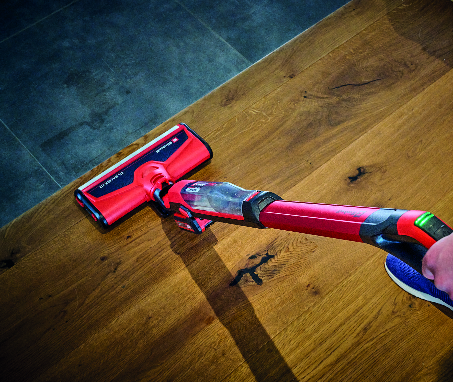 Ein roter und schwarzer Staubsauger auf Holzboden. Eine Hand hält den Staubsauger. Der Boden ist teilweise gefliest. Der Schatten ist sichtbar.
