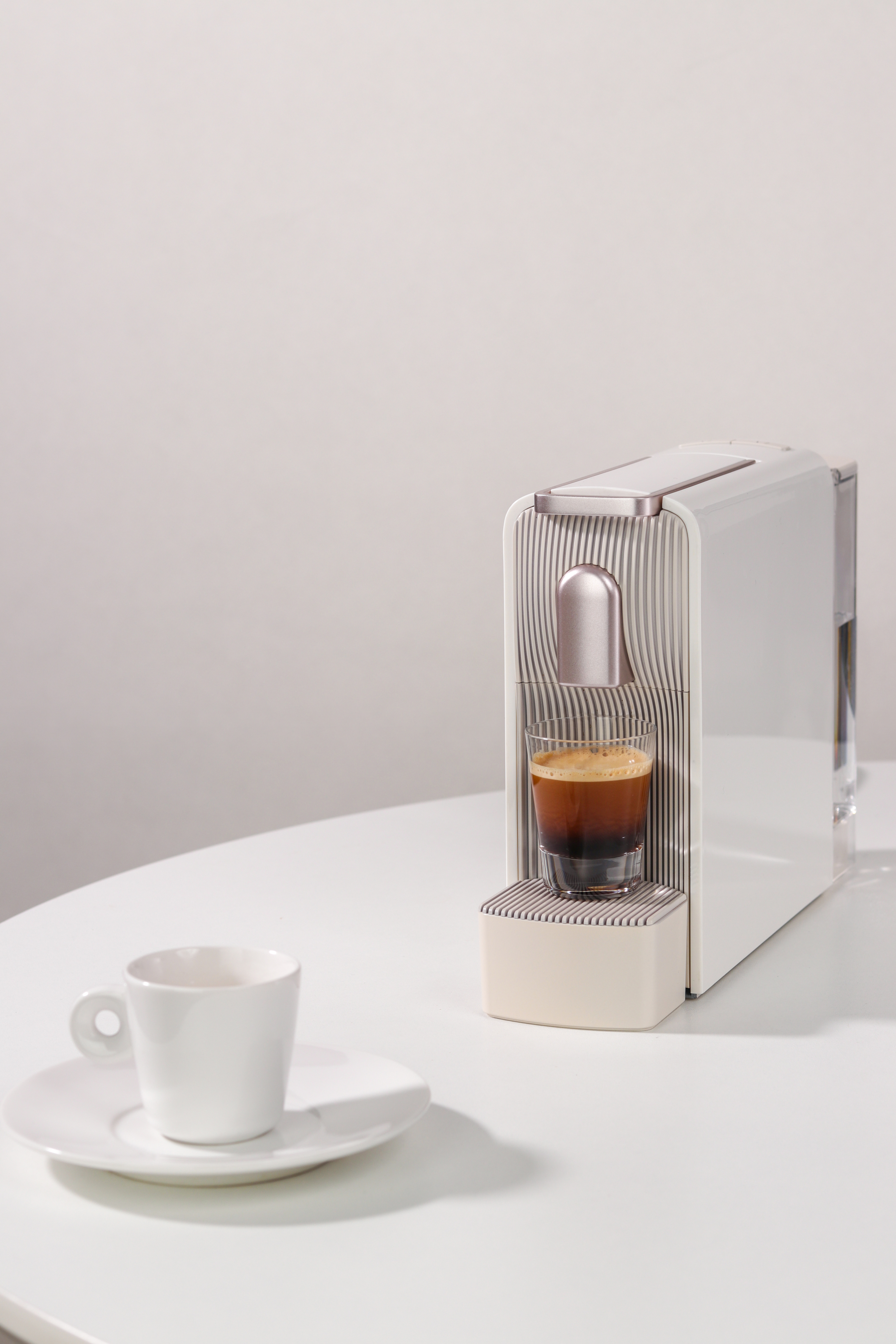 Cafetera blanca con un espresso y una taza pequeña. Todo sobre una mesa blanca.