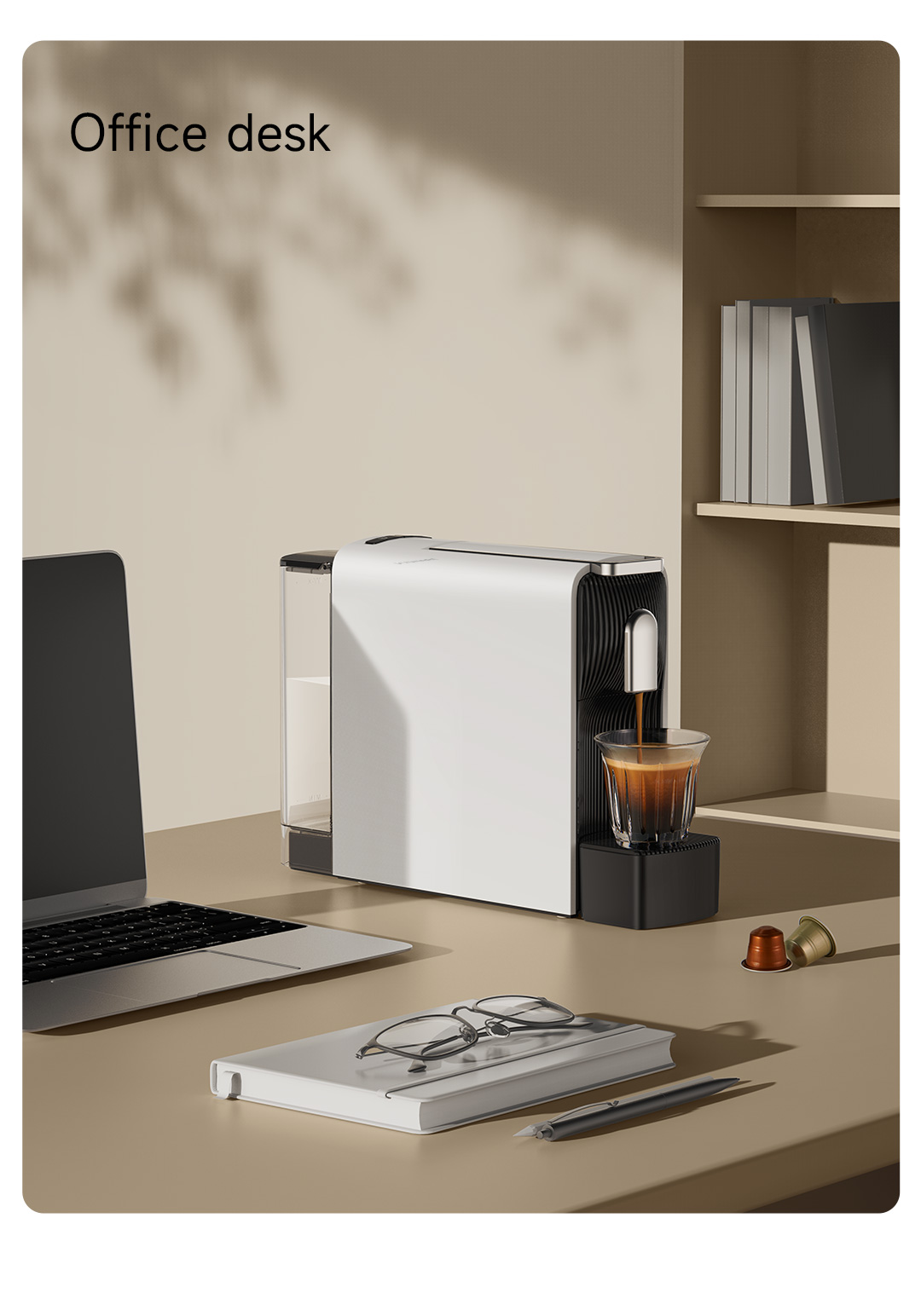 Un escritorio de oficina con cafetera, portátil, cuaderno y un vaso de café. Cápsulas en la mesa.