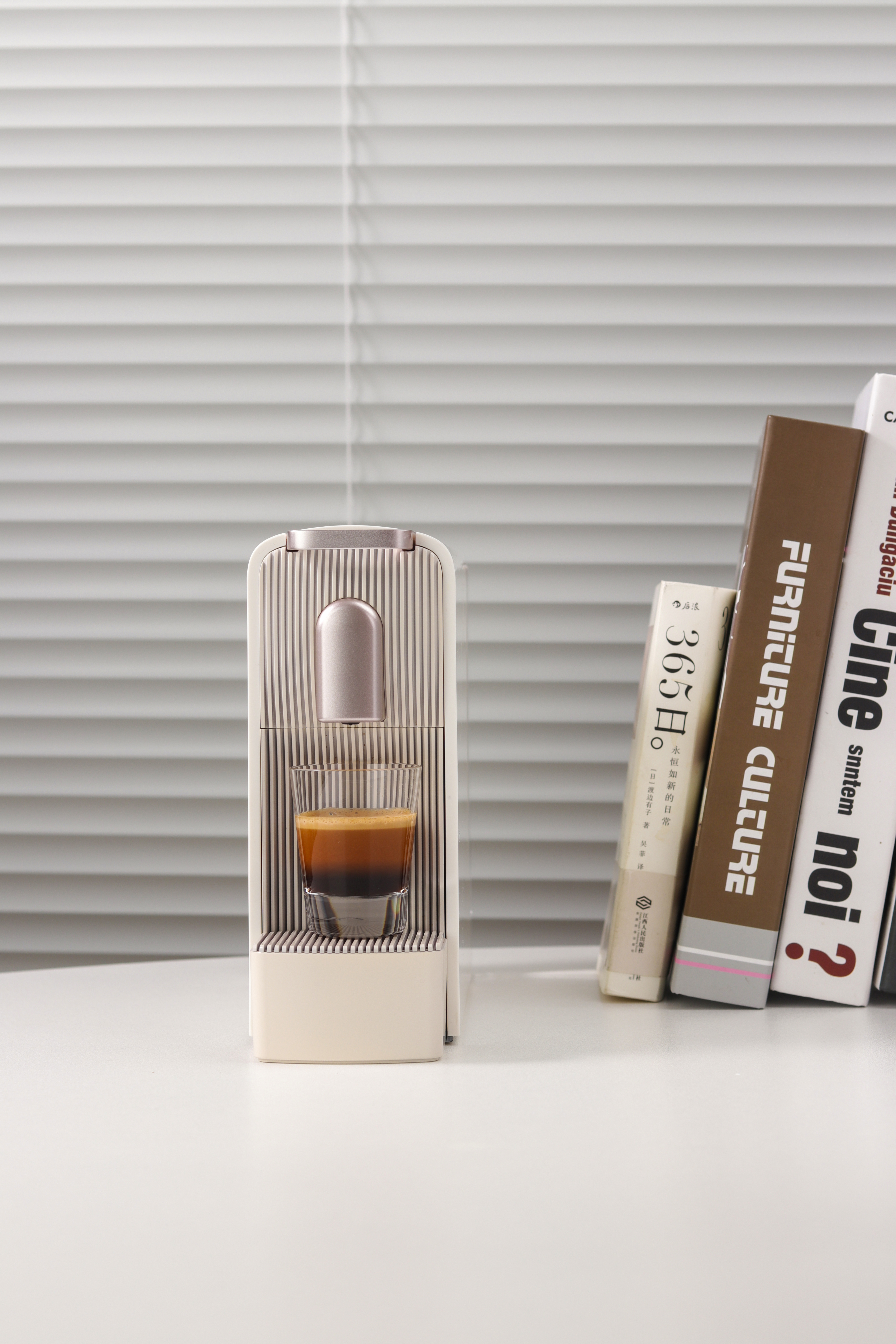 Una cafetera espresso con un vaso de café sobre una mesa blanca, con libros al fondo y persianas.