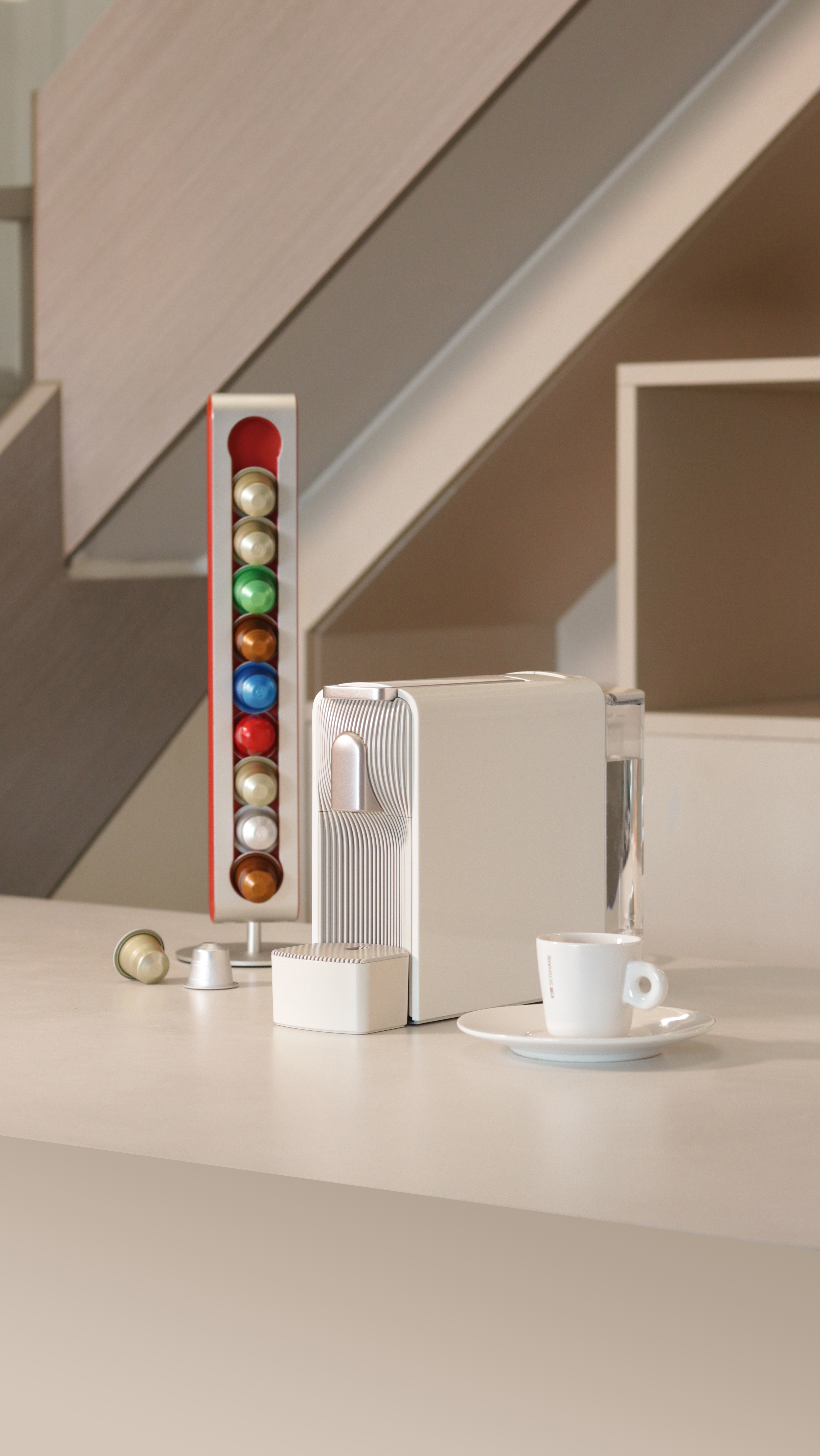 Una cafetera blanca con cápsulas, taza y platillo, y un soporte con cápsulas coloridas están sobre una superficie blanca.