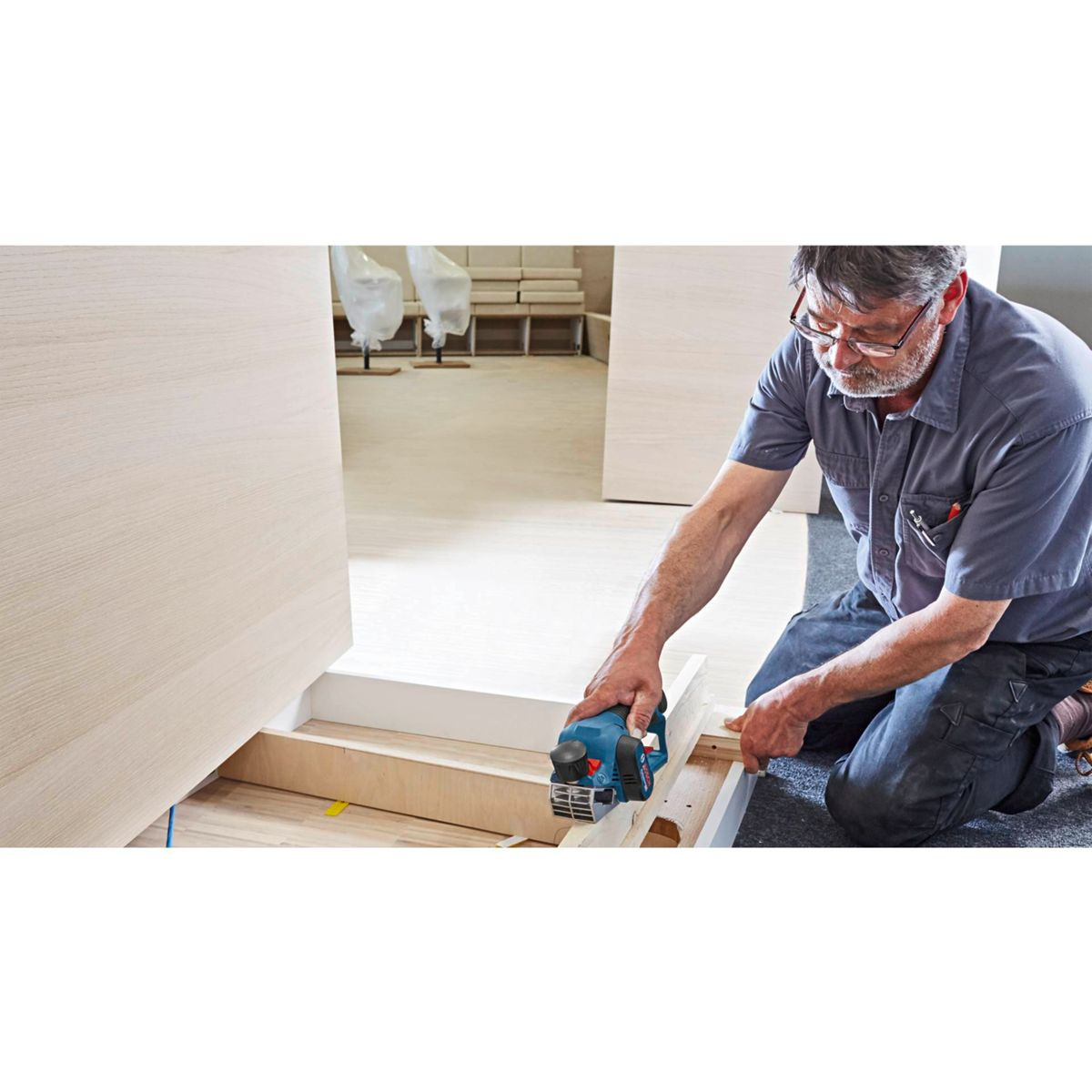 Un falegname usa una pialla elettrica blu sul legno. Si inginocchia su un tappeto grigio, lavorando su un telaio di legno. Sfondo con porte e mobili.