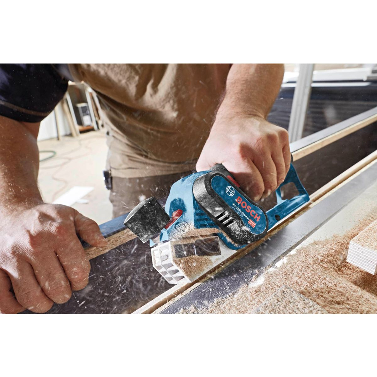 Un carpentiere pialla il legno con una pialla elettrica Bosch. Segatura vola. La pialla è blu e nera. Il carpentiere indossa una camicia marrone.
