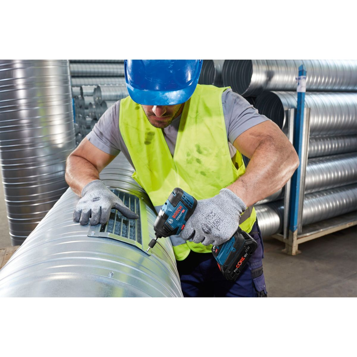 Un trabajador con casco azul y chaleco amarillo usa un taladro azul y negro en una tubería metálica. Hay guantes puestos.