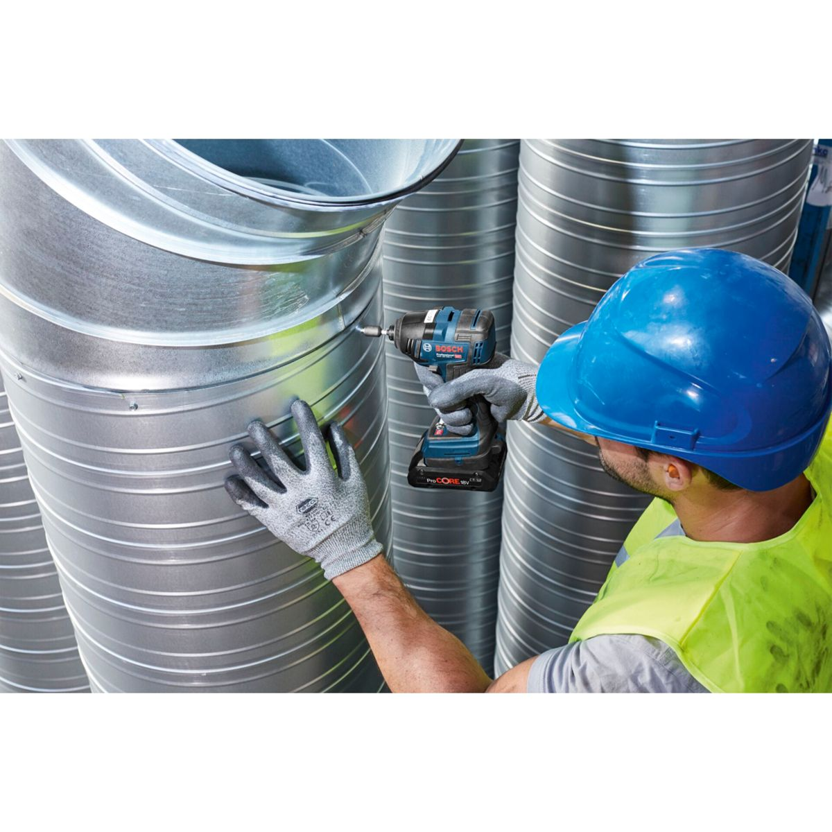 Un trabajador con casco azul y chaleco de seguridad usa un taladro Bosch en un conducto de ventilación metálico. La escena es industrial.