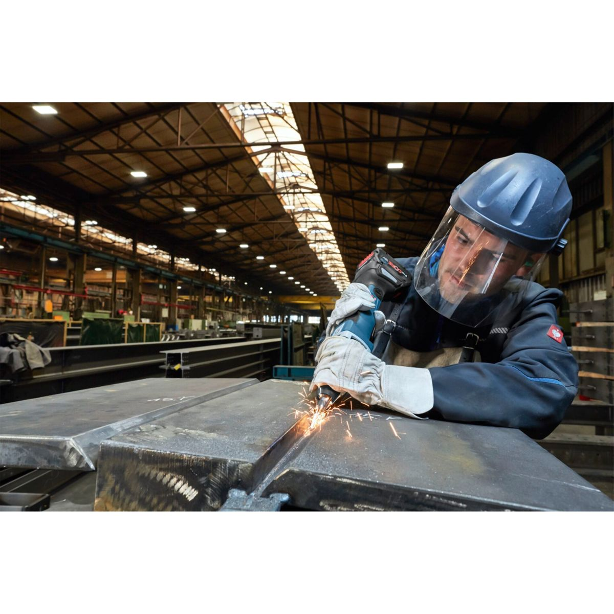 Robotnik szlifuje metal w fabryce. Iskry lecą. Nosi kask, osłonę twarzy, rękawice i odzież roboczą.