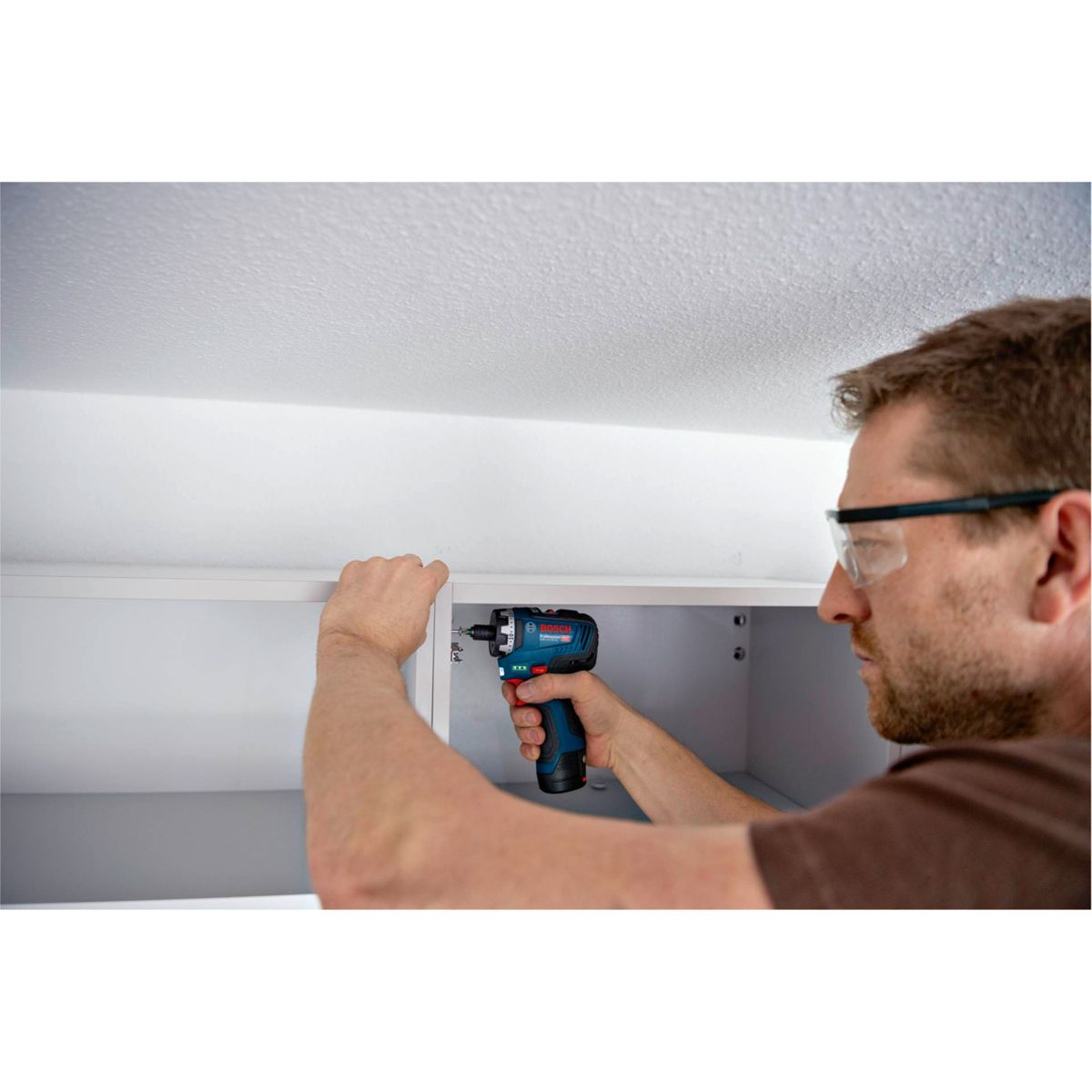 Ein Mann mit Schutzbrille benutzt einen blau-schwarzen Bohrer, um etwas in einem weißen Schrank unter einer weißen Decke zu installieren.