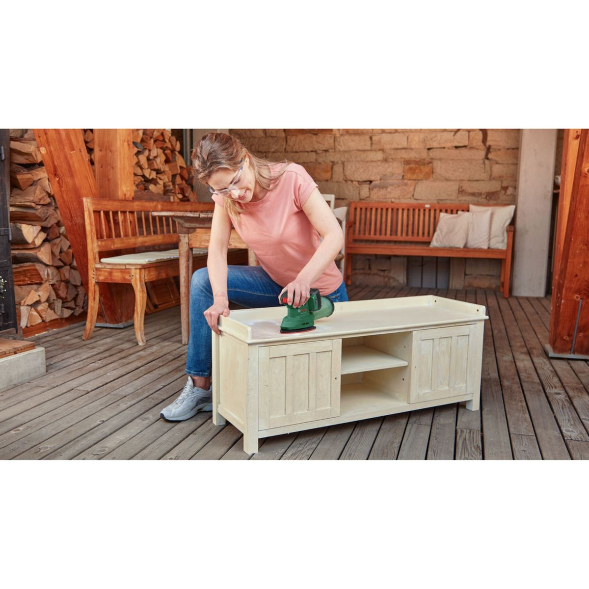Une femme ponce une armoire en bois avec une ponceuse verte. Elle porte des lunettes et un jean. Fond : bois, banc, bois de chauffage.