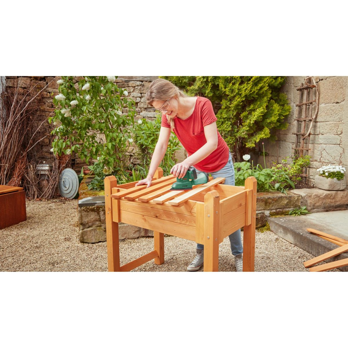 Une femme ponce des planches de bois sur un lit de jardin surélevé. Elle porte une chemise rouge, un jean et des lunettes. La scène est en extérieur.