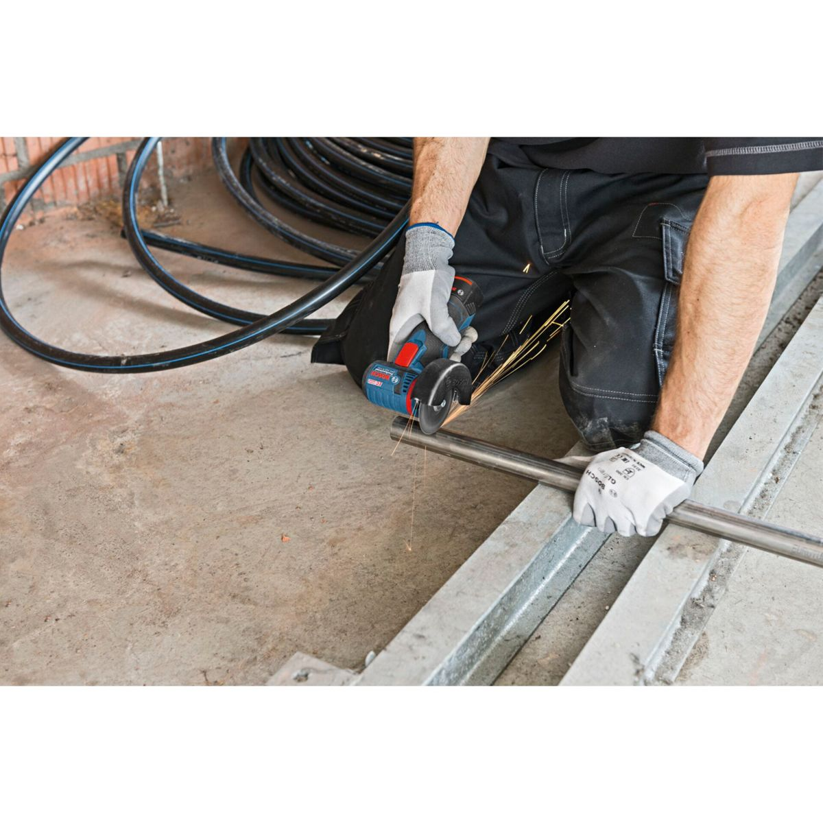 Robotnik używa szlifierki do cięcia metalowej rury. Iskry lecą, gdy narzędzie się styka. Robotnik ma rękawice i klęczy.