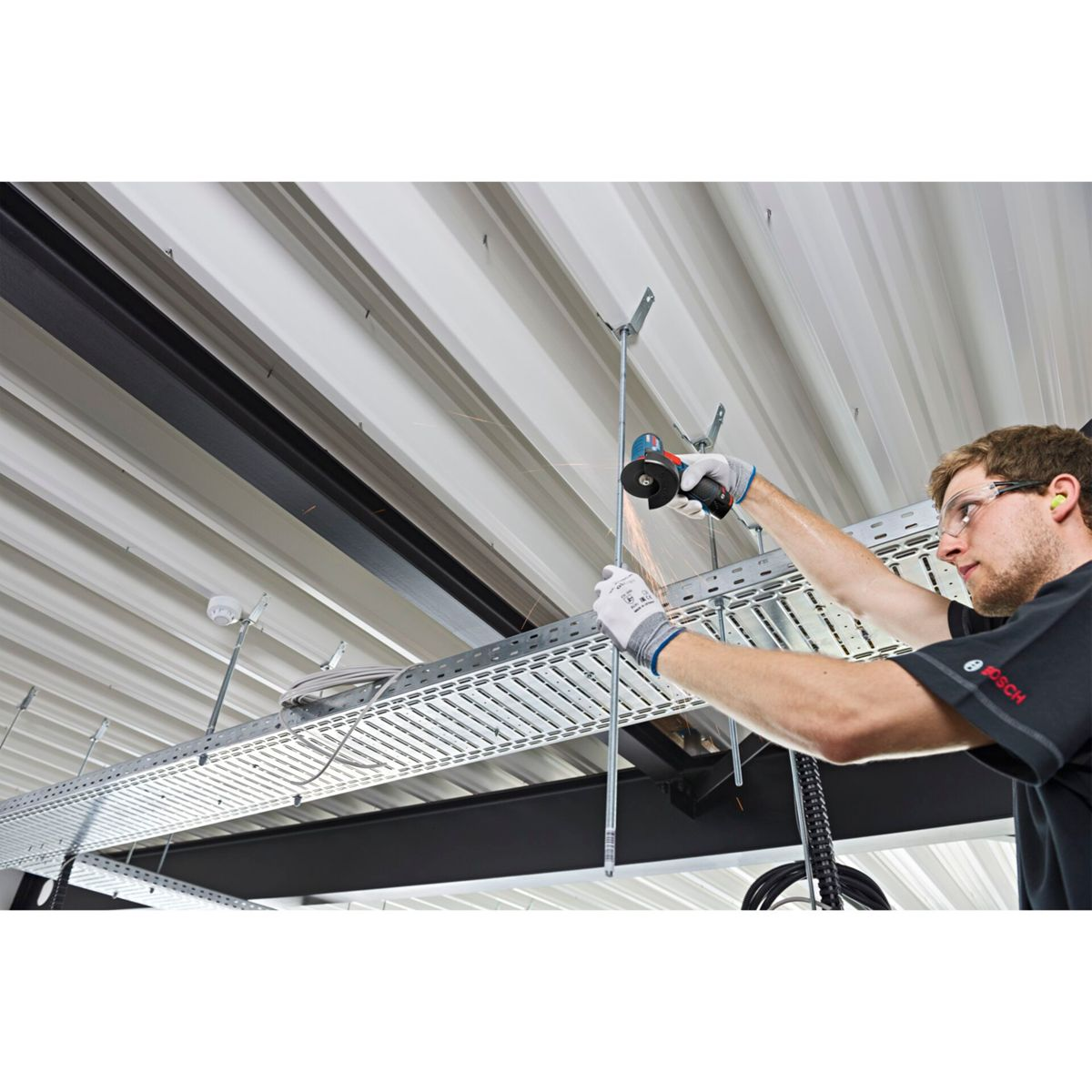 Mężczyzna używa szlifierki kątowej do cięcia metalowego pręta, lecą iskry. Powyżej, metalowy sufit z korytkami kablowymi. Noszone są okulary ochronne i rękawice.